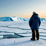 A serene, professional depiction of the Canadian Arctic. The foreground features a fully clothed indigenous elder, wearing modest, traditional winter attire, observing a vast, melting glacial landscape at sunset. Ice floes are breaking apart in the distance, and the subtle outline of a polar bear is visible on a remaining ice sheet, conveying environmental change. The scene emphasizes the vastness and vulnerability of the Arctic. Perfect anatomy, correct proportions, natural pose, well-formed hands, proper finger count, natural body proportions. Professional photography, high-resolution, safe for work, appropriate content, fully clothed, family-friendly.